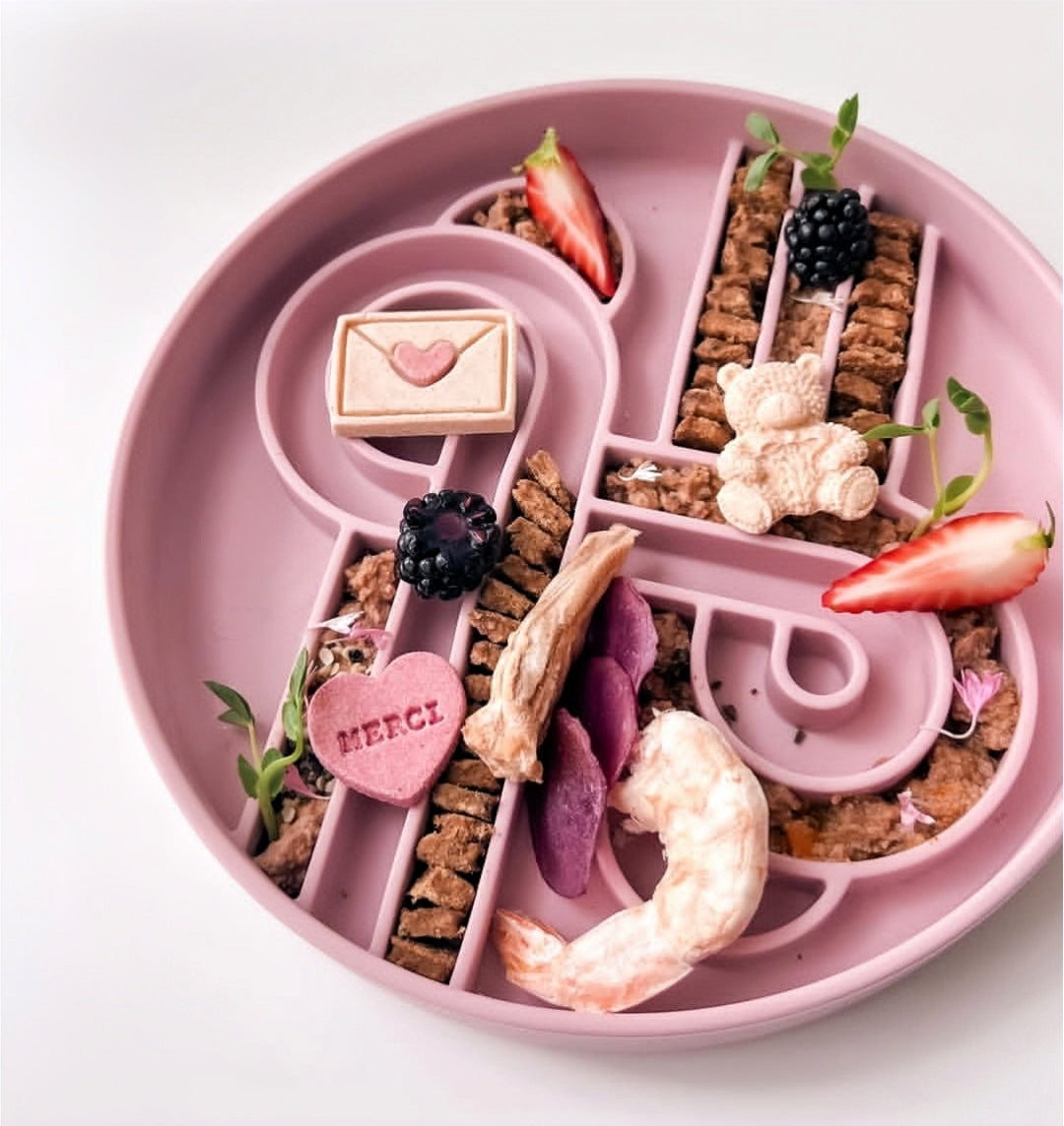 Pink Ollie b good slow feeder with divided sections containing food arranged, like freeze dried prawn in a decorative pattern on a white background. 