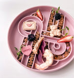 Pink Ollie b good slow feeder with divided sections containing food arranged, like freeze dried prawn in a decorative pattern on a white background. 
